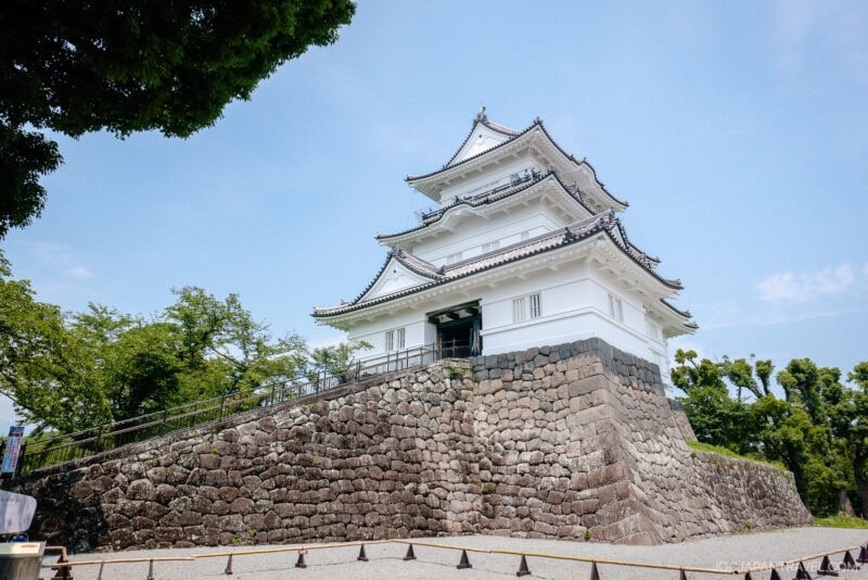 A traditional Japanese castle, like Odawara Castle, with white walls and layered roofs stands on a tall stone foundation, surrounded by trees and under a clear blue sky.