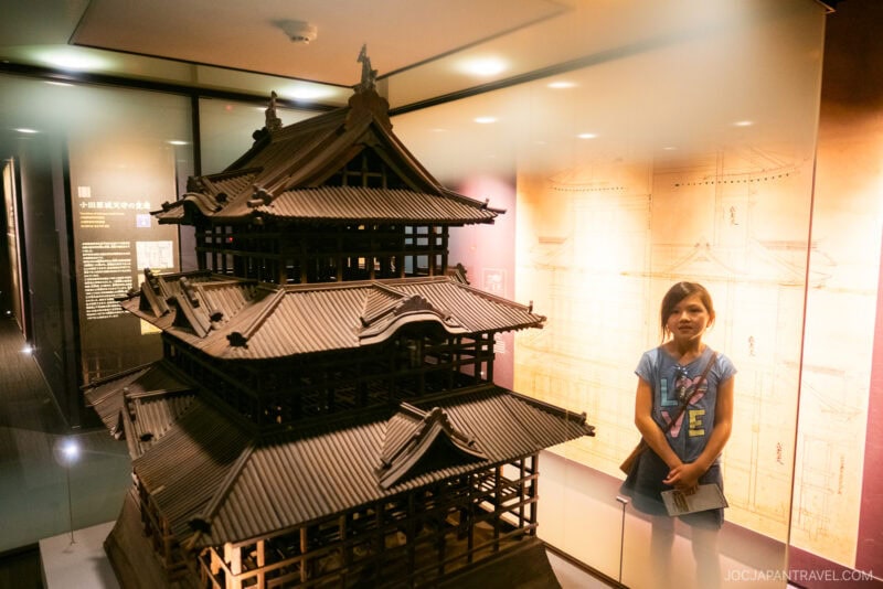 A young girl stands next to a large wooden model of Odawara Castle inside a museum, with architectural drawings displayed on the wall behind her.