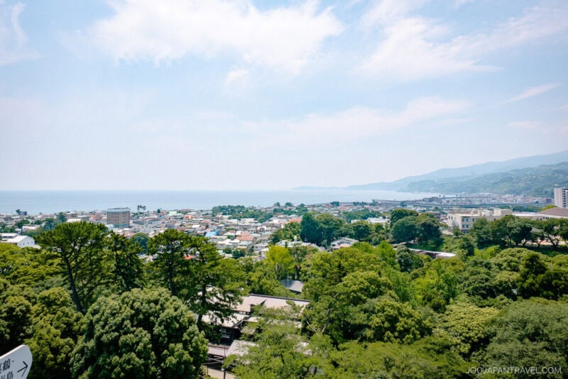 A scenic view of a coastal town with lush green trees in the foreground, buildings and houses—plus the historic Odawara Castle—in the middle, and the ocean and distant hills under a partly cloudy sky in the background.