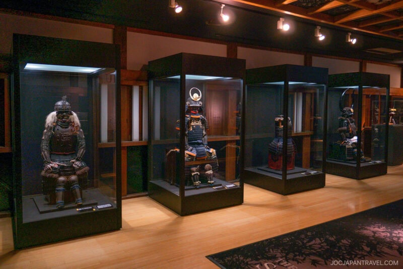 Four traditional Japanese samurai armors are displayed in glass cases in a dimly lit museum room at Odawara Castle, featuring wooden floors and ceiling beams. Each unique suit showcases intricate historical craftsmanship.