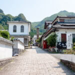 Okawachiyama (大川内山) – Village of Secret Kilns in Saga A paved stone walkway leads between traditional Japanese buildings with white walls and black tiled roofs in Okawachiyama, set against lush green mountains under a clear blue sky.