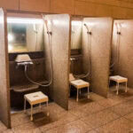 Onsen in Japan – A Guide to Hot Springs and Etiquette A row of partitioned shower stations in a Japanese bathhouse, each with a handheld showerhead, mirror, small stool, and washbowl—showing how to enjoy onsen by cleansing thoroughly in this clean, well-lit area with elegant stone surfaces.