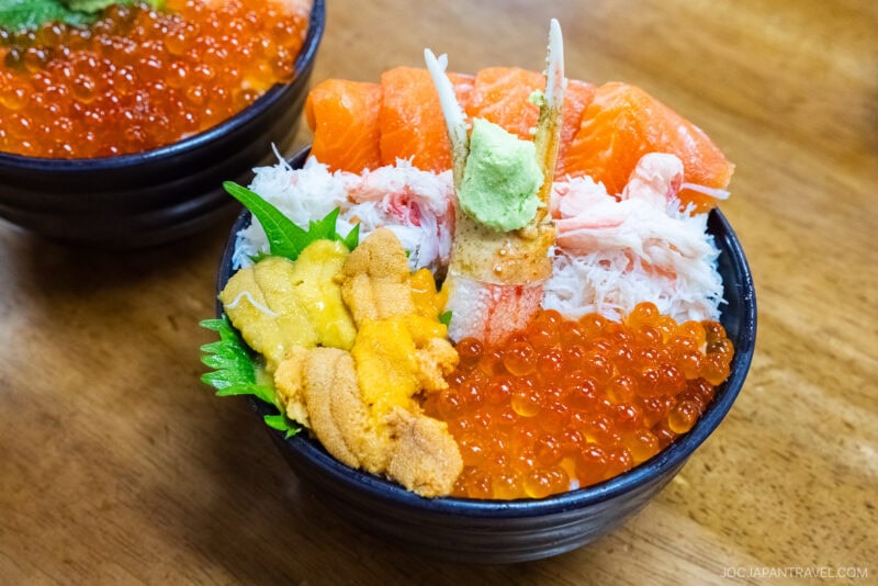 A bowl of Japanese seafood donburi inspired by Otaru, topped with salmon sashimi, crab meat, sea urchin, salmon roe, a shiso leaf, and wasabi on rice. Another similar bowl is partially visible on the wooden table.