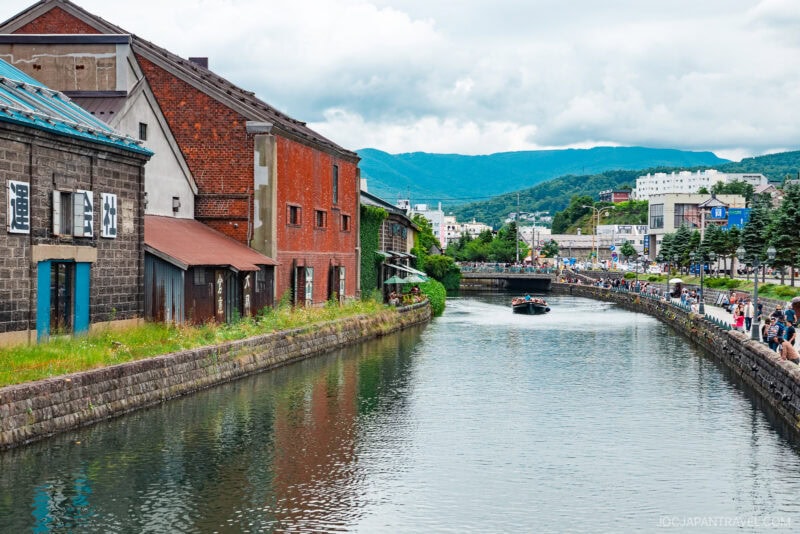 A scenic canal in Otaru lined with old warehouses and brick buildings, with people strolling along the waterfront and a boat cruising on the water, set against a backdrop of green hills and cloudy skies.