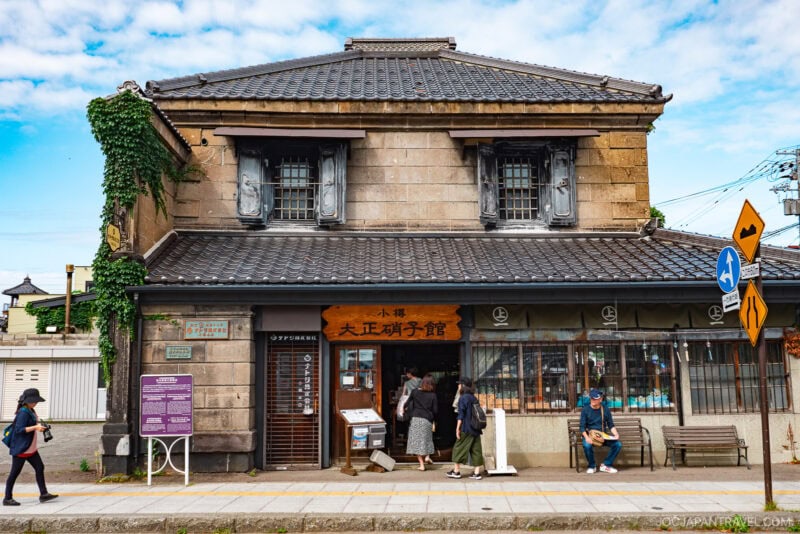 A traditional stone building in Otaru, Japan, with a tiled roof, large windows, and a wooden sign. Several people stand and sit outside, while ivy grows on the left side beneath the blue sky.