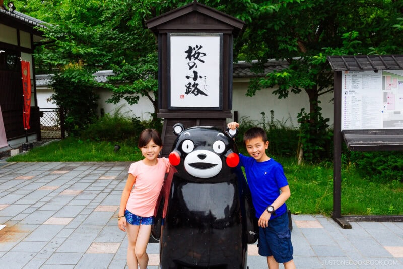 Two smiling children pose with a large black bear mascot statue at Sakuranobaba Josaien. The bear has red cheeks and a friendly face, while trees and Japanese signs appear in the background.
