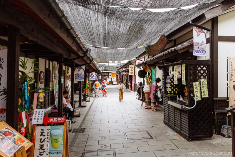 A traditional Japanese street in Sakuranobaba Josaien with shops on both sides, colorful signs, and a net canopy overhead. People in kimonos walk along the stone-paved path, creating a lively and cultural atmosphere.