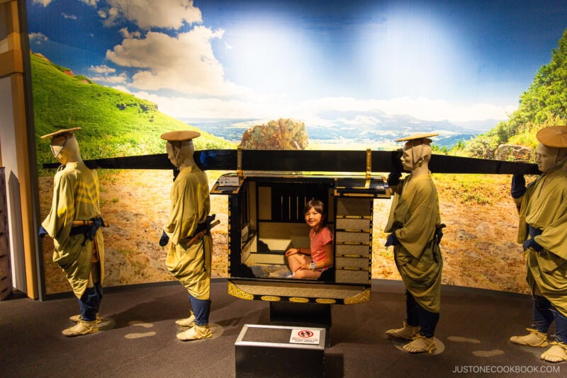 A child sits inside a traditional Japanese palanquin at Sakuranobaba Josaien, carried by mannequins dressed as Edo-period porters, with a scenic countryside backdrop.