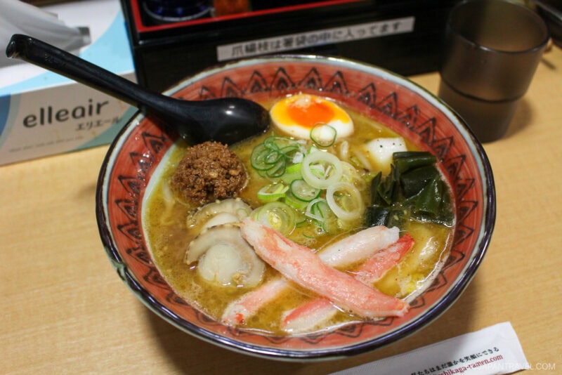 A bowl of ramen topped with sliced green onions, a soft-boiled egg, minced meat, crab sticks, seaweed, and fish cake is served with a black spoon on a wooden table—a cozy dish often featured in the Sapporo winter guide.