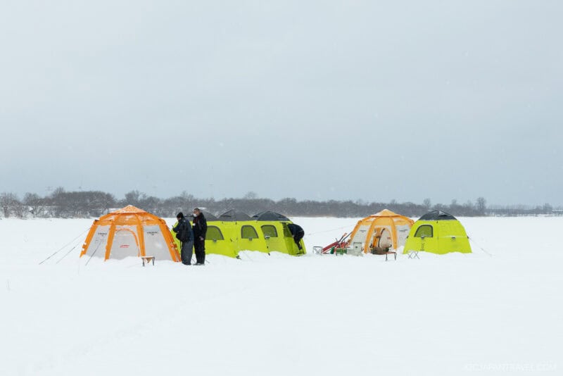 Four colorful tents (orange and green) are set up on a snowy plain. A few people, bundled in winter clothing, stand nearby under a gray sky—a scene right out of a Sapporo winter guide.