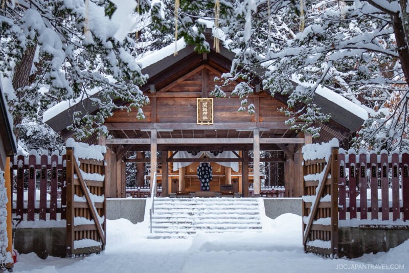 A traditional wooden Japanese shrine stands surrounded by snow-covered trees and ground, with steps leading up to the entrance and a wooden fence on either side—a serene scene fit for any Sapporo winter guide.