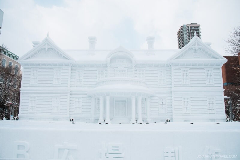 A large, detailed snow sculpture of a classical-style building with pillars and ornate features stands outdoors—a highlight in the Sapporo Winter Guide—with city buildings visible in the snowy background.