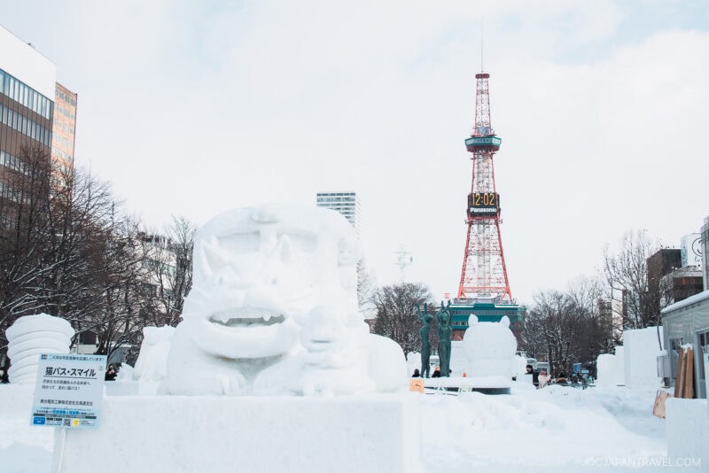 Snow sculptures and icy structures grace a snowy park in Sapporo, with the TV Tower in the background. This lively scene, featured in many a Sapporo winter guide, is alive with festivities and surrounded by city buildings.