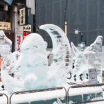 Sapporo in Winter: Festivals, Attractions & Activities Snow-covered ice sculptures, including a winged creature and a crescent moon, stand on a city street with Japanese signs and restaurants in the background—a magical scene from any Sapporo winter guide. Metal railings surround the sculptures.