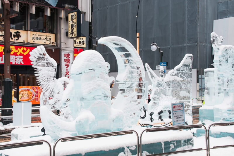 Snow-covered ice sculptures, including a winged creature and a crescent moon, stand on a city street with Japanese signs and restaurants in the background&mdash;a magical scene from any Sapporo winter guide. Metal railings surround the sculptures.