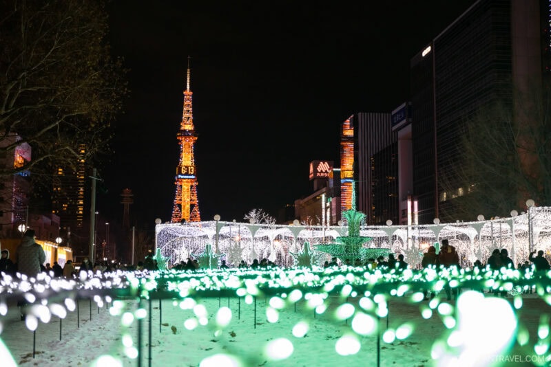 A nighttime scene of a city illuminated by festive lights, with a brightly lit tower in the background and crowds enjoying glowing decorations on snowy ground—a magical moment featured in any Sapporo winter guide.