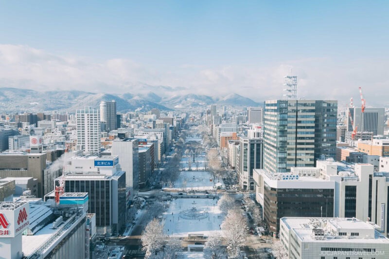 A snowy aerial view of Odori Park in the center of Sapporo, Japan, surrounded by tall buildings and distant mountains under a light blue sky—a perfect scene for any sapporo winter guide.