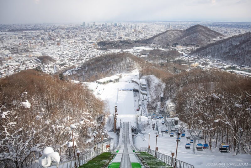A snowy ski jump ramp leads down to a stadium, surrounded by snow-covered trees and hills, with a cityscape and distant mountains visible under a cloudy sky—perfect for any Sapporo winter guide.