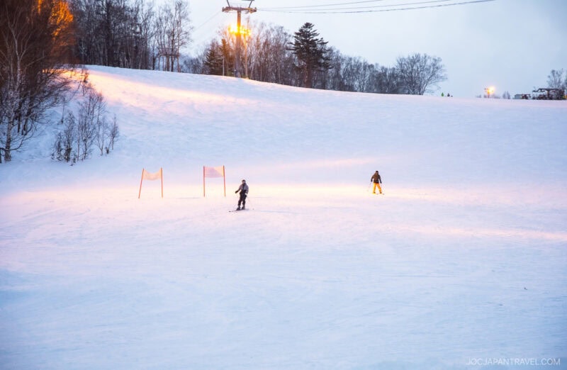 Two skiers glide down a snow-covered slope at dusk, illuminated by soft yellow lights. Bare trees frame the scene beneath an overcast sky. This peaceful moment could be straight from a Sapporo winter guide.
