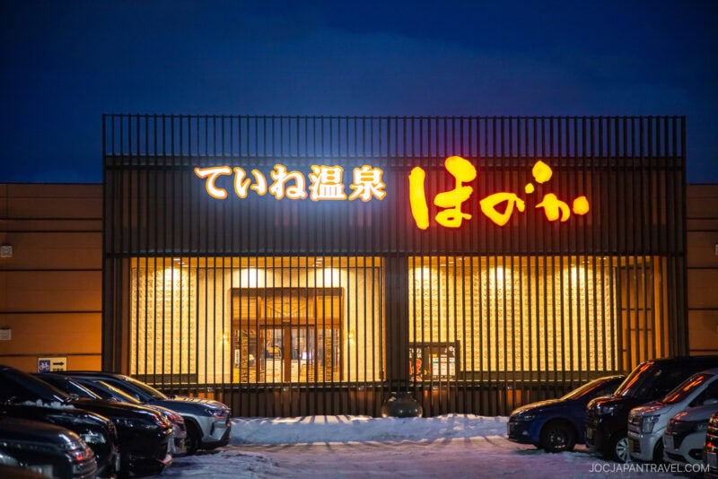 A Japanese onsen building at night with illuminated signs in Japanese above the entrance. Snow covers the ground and several parked cars line the front, offering a classic scene for any Sapporo winter guide.