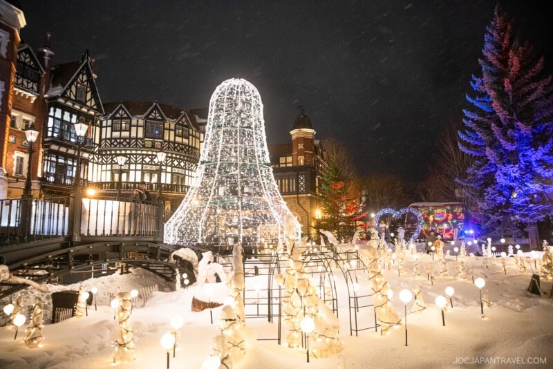 A snowy outdoor scene at night features illuminated buildings, a large cone-shaped light display, and white trees decorated with glowing lights, capturing the magic highlighted in any Sapporo winter guide.