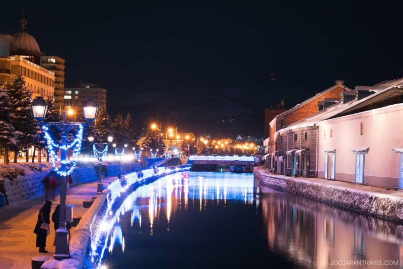 A snowy canal at night is lined with illuminated street lamps and buildings, their lights reflecting on the calm water. Blue and white festive lights decorate the trees and railing, creating a magical scene straight from a Sapporo winter guide.