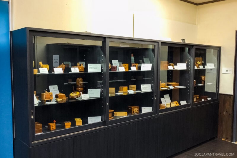 Three glass display cases filled with various wooden puzzles and objects, each labeled with small name cards, are situated against a yellow-beige wall in the Sekisho Karakuri Museum exhibition setting.