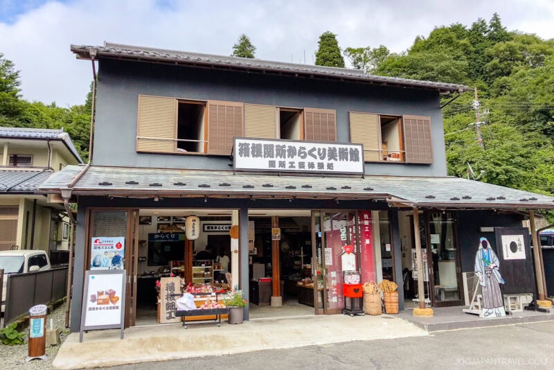 A traditional two-story Japanese building with open windows and signs in Japanese, housing a shop selling local goods near the Sekisho Karakuri Museum. The entrance displays souvenirs and a life-size cutout, with trees visible in the background.