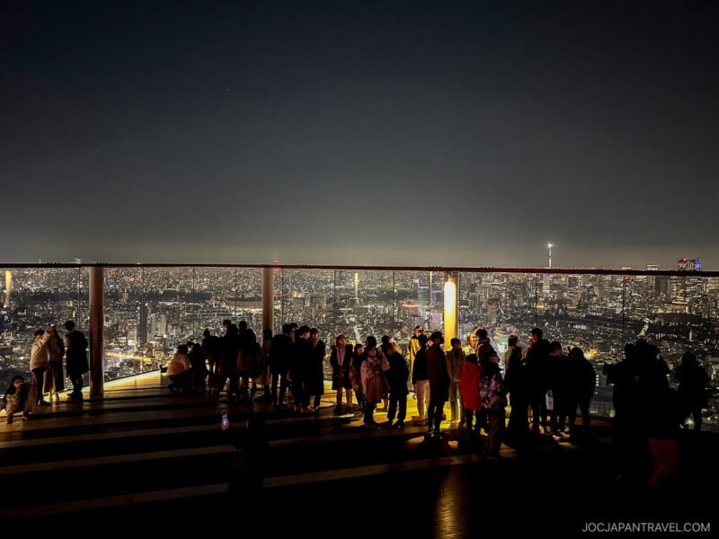 A group of people stand on the Shibuya Sky rooftop observation deck at night, overlooking a brightly lit cityscape with countless buildings and lights stretching into the distance.