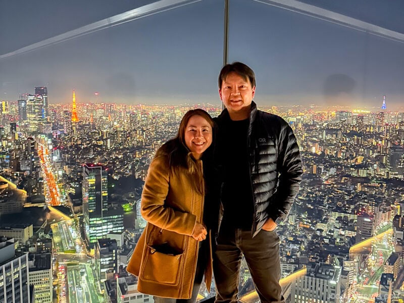 A smiling couple stands close together inside the glass-walled Shibuya Sky observation deck at night, with a brightly lit cityscape and towering skyscrapers stretching out behind them.
