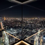 Shibuya Sky (渋谷スカイ) – Panoramic Views Above Shibuya Crossing A nighttime cityscape of Tokyo viewed from a high-rise observation deck like Shibuya Sky, with bright city lights, dense buildings, and reflections on tall glass windows forming a striking V-shaped perspective.