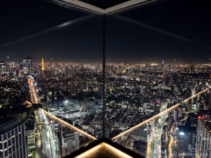 A nighttime cityscape of Tokyo viewed from a high-rise observation deck like Shibuya Sky, with bright city lights, dense buildings, and reflections on tall glass windows forming a striking V-shaped perspective.