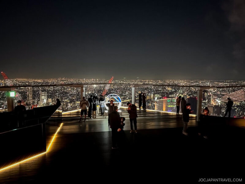 People stand on the Shibuya Sky rooftop observation deck at night, overlooking a brightly lit cityscape that stretches into the distance. Some take photos as the glowing city lights create a vibrant backdrop under the dark sky.