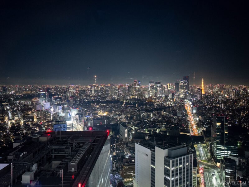 Aerial view of a city at night with many illuminated buildings, busy streets with car lights, and a dense urban skyline stretching into the distance under a dark sky, reminiscent of the stunning perspective from Shibuya Sky.