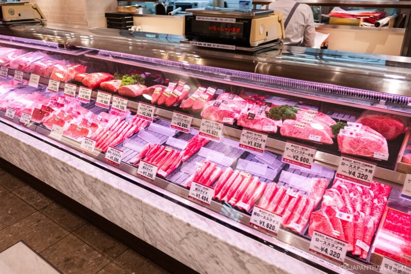 A refrigerated display case on the Isetan Food Floor in a Japanese supermarket showcases assorted cuts of raw beef, neatly arranged with price and description tags under bright lighting. A person is seen working behind the counter.