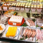 Shinjuku ISETAN Food Floor – Tokyo’s Gourmet Depachika Experience A display case in a Japanese market, reminiscent of the Isetan food floor, features packaged seafood, fish fillets, yellow tamagoyaki, jars of condiments, and labeled price tags in Japanese.