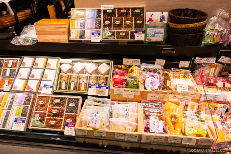 A store display of assorted Japanese packaged foods and sweets, neatly arranged in boxes and baskets with labels and price tags, reminiscent of the isetan food floor inside a well-lit shop.