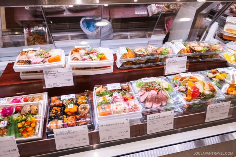 A display case on the Isetan food floor filled with various Japanese bento boxes, featuring sushi, rice dishes, vegetables, and meat, all neatly arranged and labeled with prices in a store.