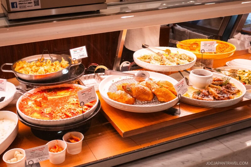 A display case on the Isetan food floor showcases various prepared Japanese dishes—curry, fried foods, noodles, and rice—each with a description card and smaller sample portions in cups in front.