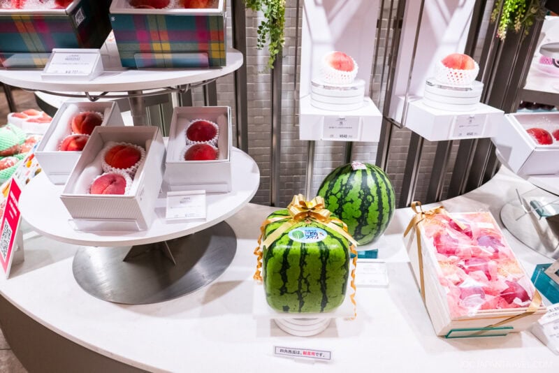 A display of luxury fruits on the Isetan food floor features a square watermelon with a gold ribbon, round melons, apples in protective foam nets, and elegant fruit gift boxes, all arranged neatly in the bright store.