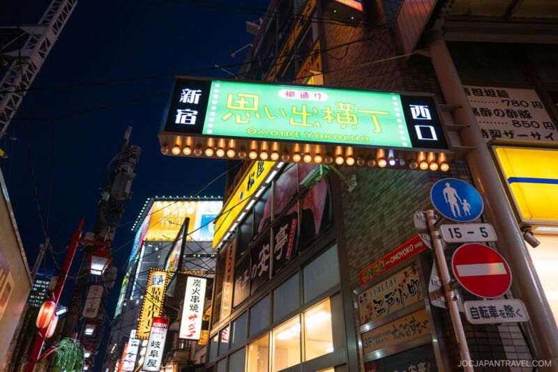 A bright neon sign for Omoide Yokocho—also known as Memory Lane—in Shinjuku, Tokyo, glows at night, surrounded by illuminated shop signs and storefronts. Japanese characters and street signs create a lively nighttime scene.
