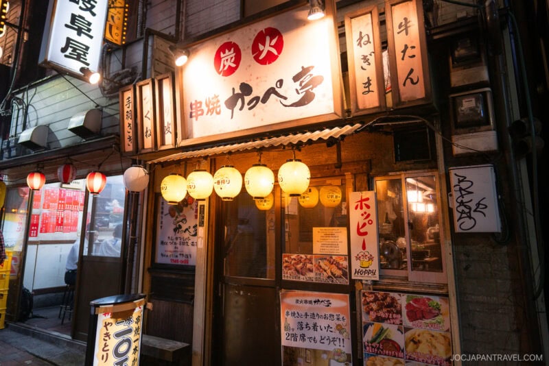A cozy Japanese izakaya with glowing paper lanterns and wooden signs evokes a stroll down memory lane, featuring handwritten menus and posters outside, inviting guests to dine in a warm, traditional setting at night.