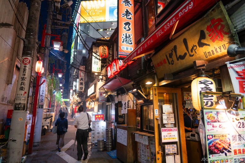 A lively street in Japan at dusk feels like memory lane, lined with brightly lit signs and cozy restaurants. People stroll along the narrow sidewalk, passing colorful storefronts and glowing lanterns.