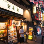 A lively alley in Japan at night, often called memory lane, features illuminated signs, restaurant menus with food and drink images, and people walking under colorful neon lights.