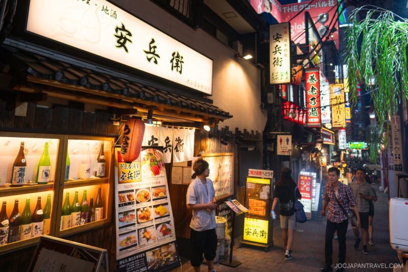 A lively alley in Japan at night, often called memory lane, features illuminated signs, restaurant menus with food and drink images, and people walking under colorful neon lights.