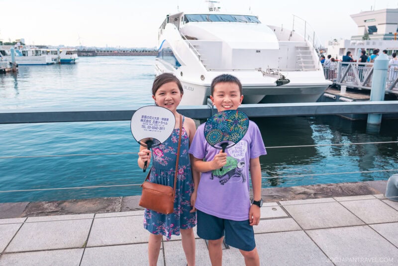 Two smiling children stand by a harbor holding round fans, as if ready for a hanabi guide, with boats docked behind them. One wears a purple shirt and shorts, the other a blue dress. A large white ship is docked in the background.