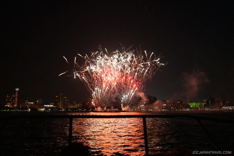 Colorful hanabi fireworks burst over a city skyline at night, reflecting on the water below. The scene is viewed from a boat, with parts of the railing visible in the foreground—an inspiring sight for any hanabi guide.