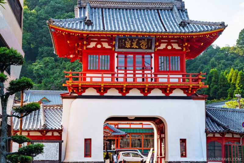 A traditional Japanese gate with ornate red and white woodwork and a tiled roof stands at the entrance to a historic Takeo Onsen building, surrounded by lush greenery and trees.