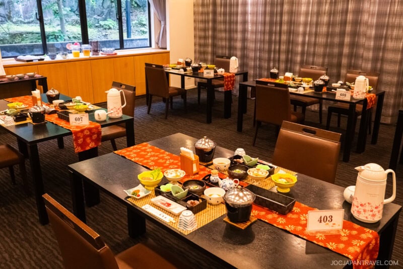A dining room at Takeo Onsen with several tables set for a Japanese meal, featuring various dishes, teapots, and tableware. Numbered signs sit on each table. Large windows reveal greenery outside, adding to the cozy, inviting atmosphere.