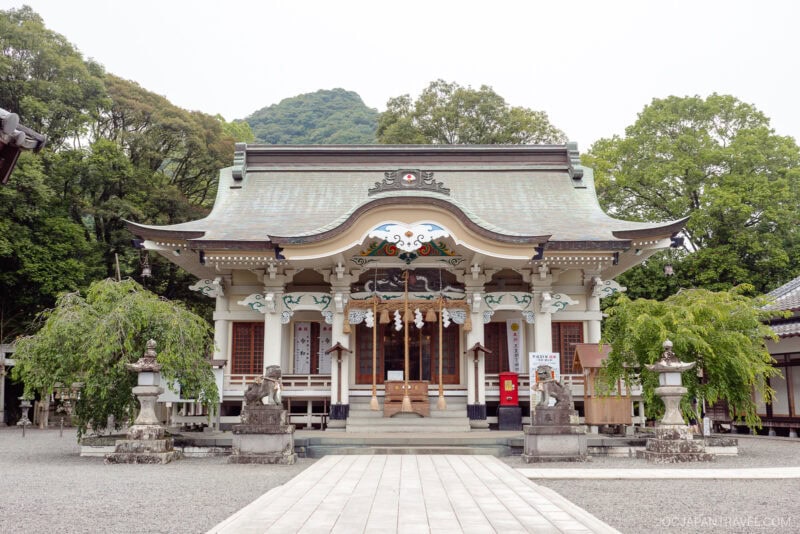 A traditional Japanese Shinto shrine with ornate architectural details, guarded by stone lion statues, nestled near Takeo Onsen, surrounded by trees and a gravel courtyard, with a mountain visible in the background.